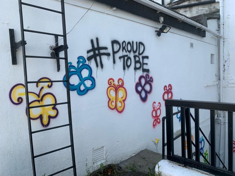Graffiti shows the words #proudtobe, surrounded by spray painted flowers 