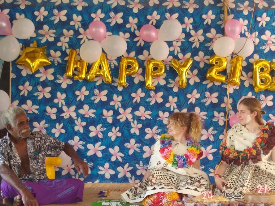 Two students sit with their host father in Fiji, surrounded by balloons and happy birthday signs