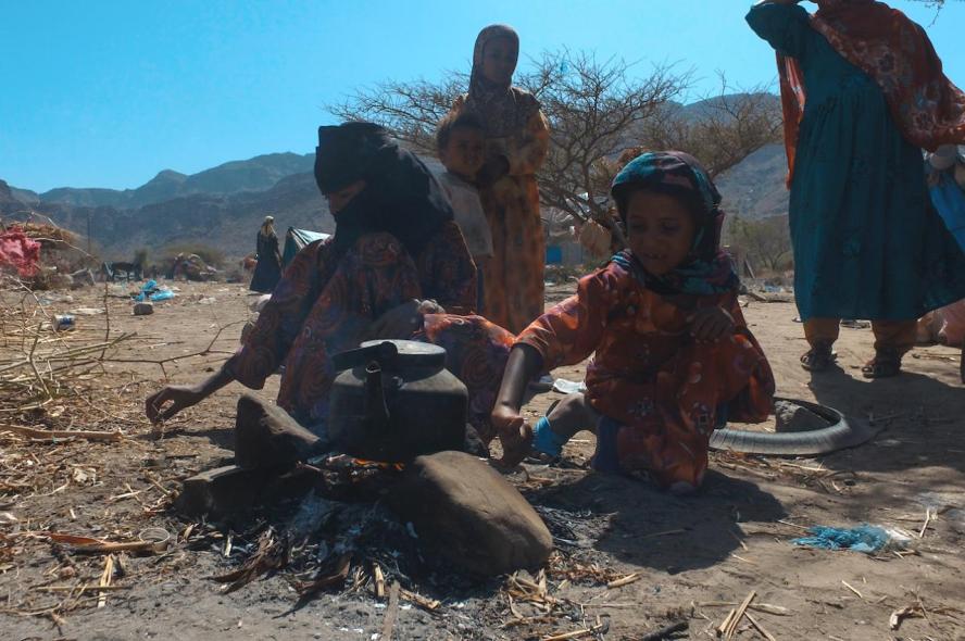 child and adult around pot on small fire on dirt ground against blue sky