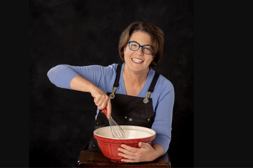 Smiling in glasses, blue shirt, and black apron holds a red mixing bowl and whisk