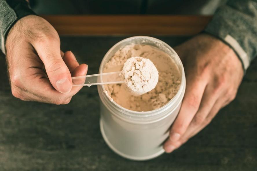 hands hold a scoop of protein powder above a countainer on a counter