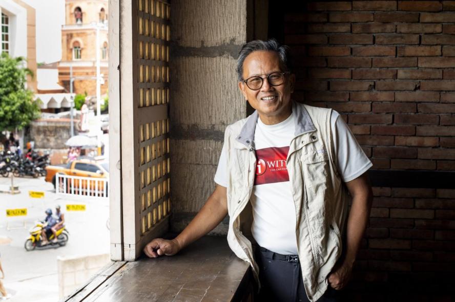 A smiling man in a vest, T-shirt and glasses stands by an open window looking onto a street