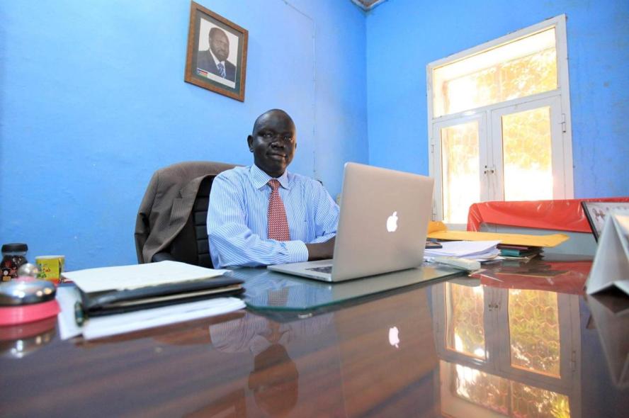 Man in blue shirt sits in office with blue walls behind a laptop at a desk