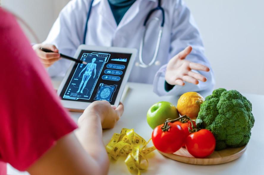 person in red shirt holds tablet with schematic of the body as doctor in white coat gestures next to plate of colorful veggies and fruits