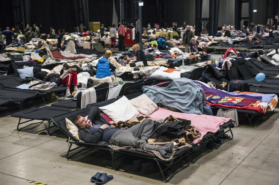 Dozens of people lying on cots with blankets in a large room with a bare floor.