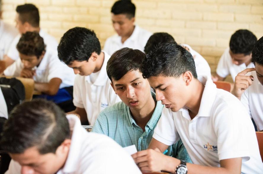 A college student in a blue collared shirt assists a younger student, in a classroom full of students in white uniform shirts.