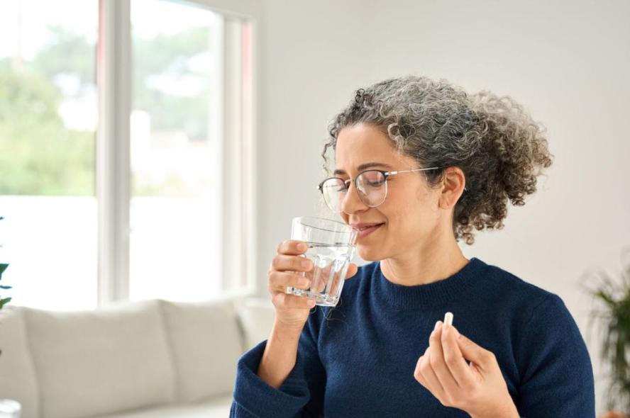 A woman with curly, graying hair takes a sip of water in preparation to swallow a large white vitamin pill
