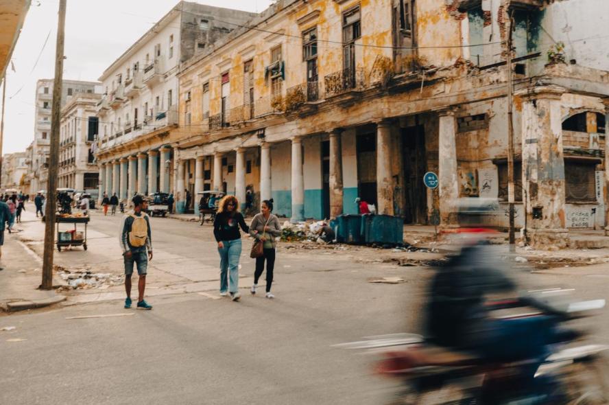 A street scene in Havana, Cuba, with a motorcycle speeding past and pedestrians walking in the street, with building facades looking distressed and trash puled up by a gutter