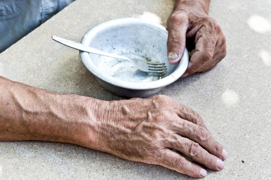 A close-up photograph of an older person's hands on a table, gripping a bowl that's empty of food but has a fork in it.