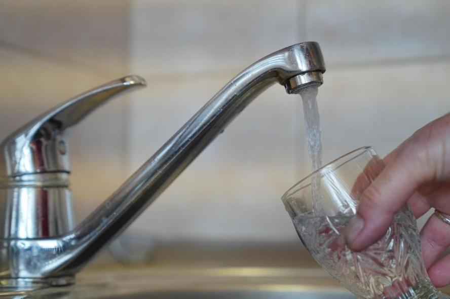 A close-up photograph shows a hand holding a drinking glass as it is filled with water from a kitchen sink faucet 