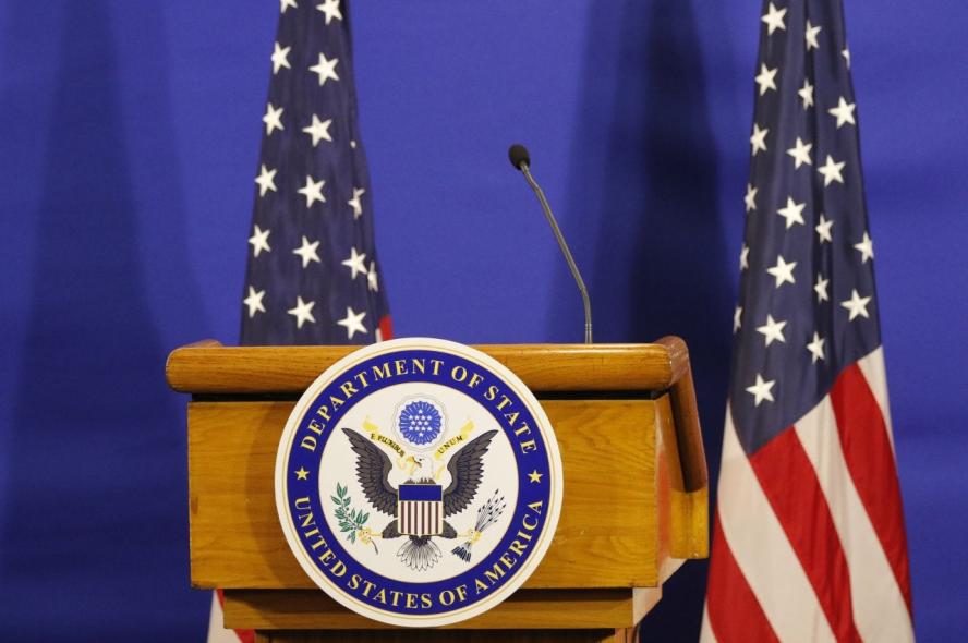 A podium with a U.S. State Department seal on the front and two U.S. flags beside it