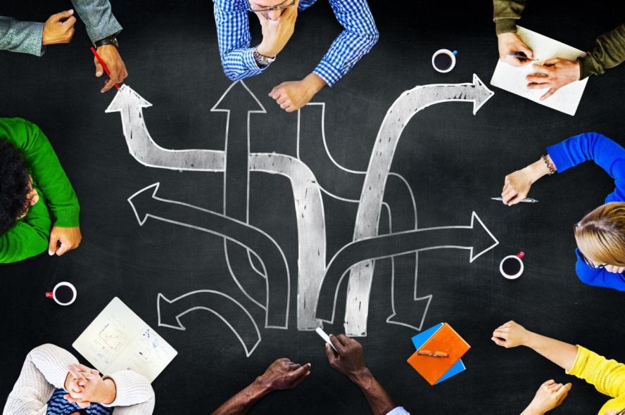 Eight people sit around a chalkboard table showing a maze of paths on its surface