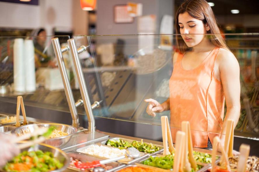 A young woman in an orange shirt chooses vegetables at a dining hall