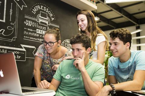 Students gather around a computer monitor. A chalkboard in the background has words written in Portuguese.
