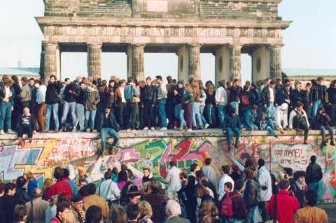 People standing on the ground and on the Berlin Wall, with a towering old building in the background. The fall of the Berlin Wall in 1989 led to German reunification because diplomats seized on the momentum after East Germany eased travel restrictions.