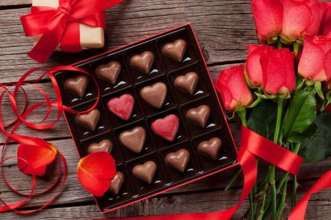 A box of heart shaped chocolates on a wooden tabletop surrounded by red ribbons and red flowers