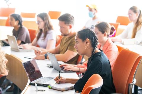 Woman with braids and black shirt sits in orange chair writing notes in a bright classroom with other students