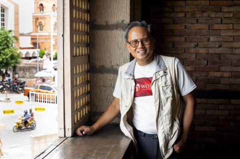 A smiling man in a vest, T-shirt and glasses stands by an open window looking onto a street