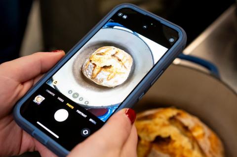 Hands hold an iphone that&#039;s taking a picture of a round loaf of bread in a bowl