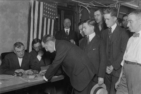 A group of men in 1917 line up to register for the draft at a desk with a flag flying behind them. A study of the voting records on conscription of U.S. legislators with draft-age sons in the 20th century shows how self-interest ruled votes