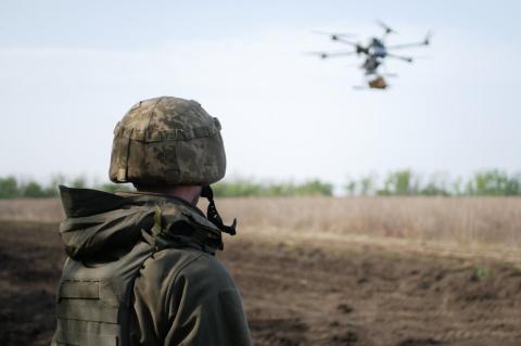 A soldier on a barren field looks up at a drone moving across the sky. Drones started as ways to gather intel on enemies, and now are used to deliver deadly attacks, as battlefields continue to evolve in Ukraine and beyond