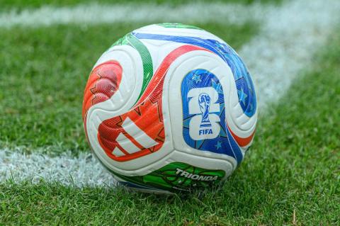 A multicolored soccer ball emblazoned with the 2026 FIFA logo sits on a green soccer field