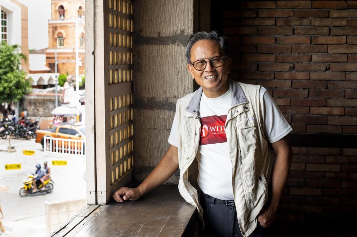 A smiling man in a vest, T-shirt and glasses stands by an open window looking onto a street