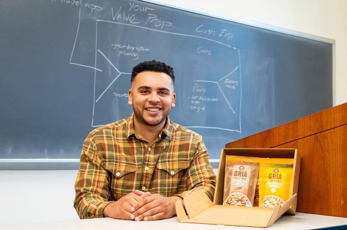 Joshua Reed-Diawuouh sits in front of a chalk board with bags of his cashew snacks at his side.