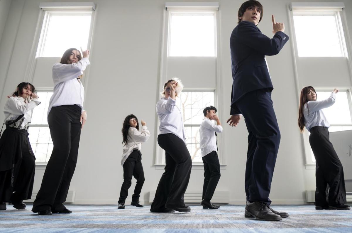 A group of people stand in a formation, dancing in a room with a white wall and windows.