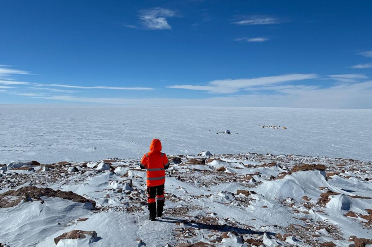 A person in a red parka stands on an outcrop of rocks looking over a glacier, with a small tent in the distance
