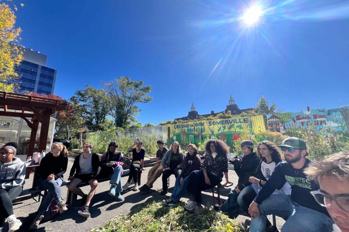A group of students seated at a farm. The words &quot;Groundworks Somerville&quot; and &quot;South Street Farm&quot; are on the wall behind them. It&#039;s a sunny, bright day.